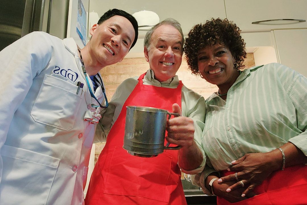 Three smiling adults—Asian dentist in lab coat, man in apron holding sifter, Black woman—indoor kitchen setting, eye-level view.